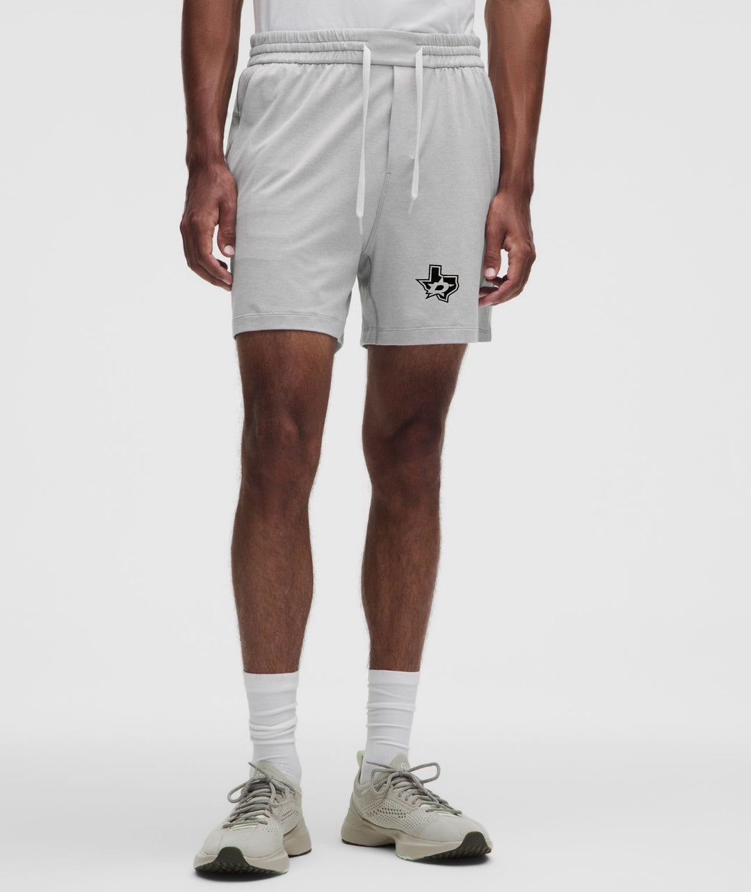 A model from the waist down wearing light gray athletic drawstring shorts with a black Texas-shaped logo on the left leg, paired with white socks and beige sneakers, against a light gray studio background.