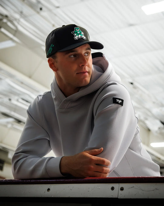 Jake Oettinger wearing a light gray hoodie and a black baseball cap with a otter logo, leaning on a bench in a rink setting.