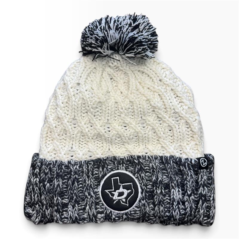 A knit pom-pom beanie hat with a cream cable-knit upper section and a dark gray and white marled lower cuff. A circular black patch with a white Texas outline and central star is stitched on the front of the cuff.