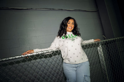 Woman wearing a white sweatshirt with text and graphics, standing against a chain-link fence.