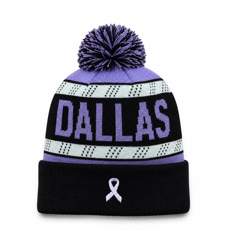A purple and black knit beanie with a pom-pom, featuring the word 'DALLAS' in purple on a black band, a white patterned stripe, and a small lavender cancer awareness ribbon on the cuff.