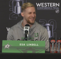 Esa Lindell speaking at a press conference with branding and logos in the background