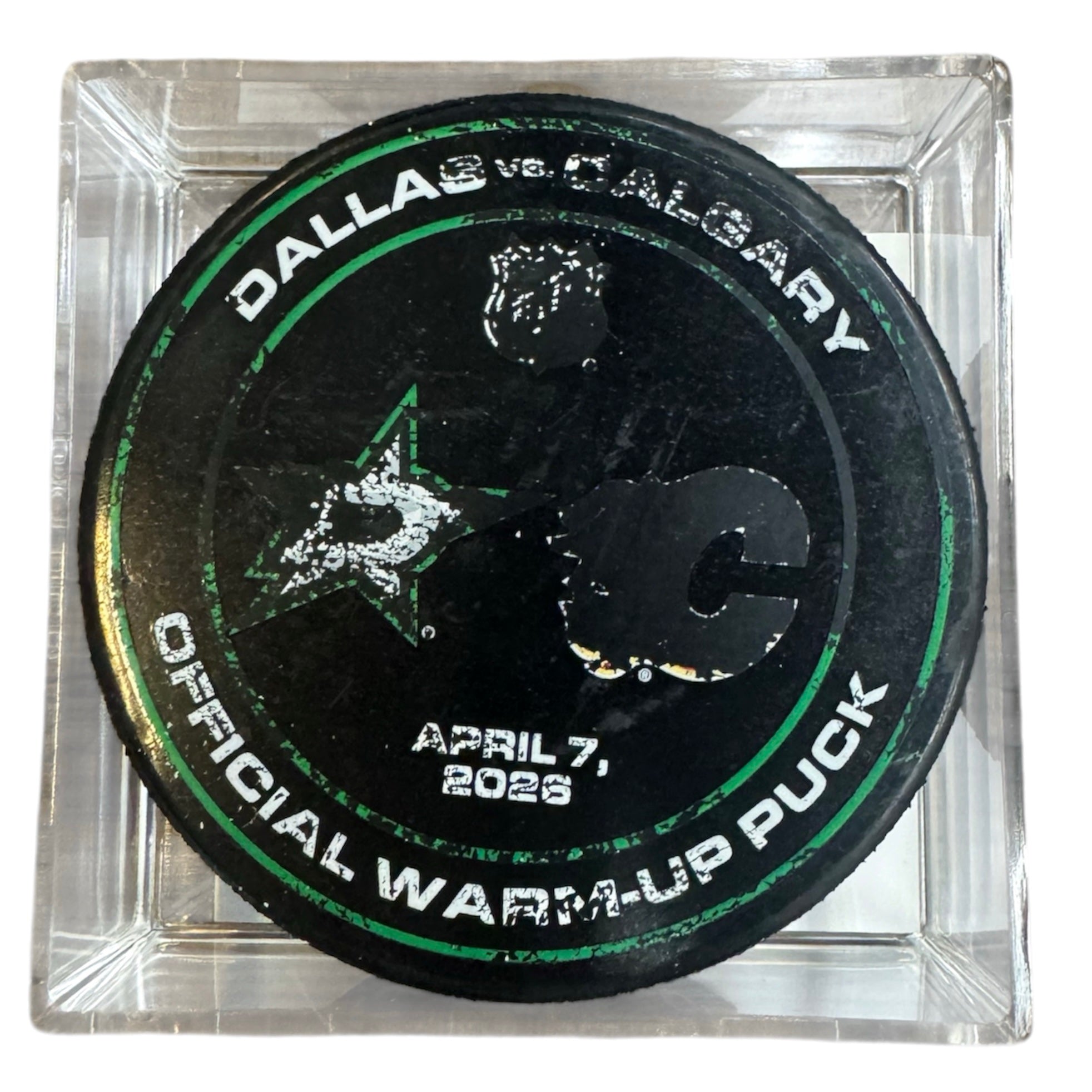 A black Dallas vs Calgary official warm-up hockey puck displayed inside a clear acrylic block. The puck features white and green text and logos, including the NHL shield and team logos, dated April 7, 2026.